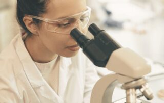 Laborantka w białym fartuchu i okularach ochronnych obserwuje preparat pod mikroskopem w laboratorium, podczas analizy próbki nasienia.