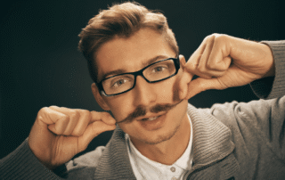 Młody mężczyzna w okularach podkręcający wąsy, symbol kampanii Movember promującej profilaktykę zdrowia mężczyzn.